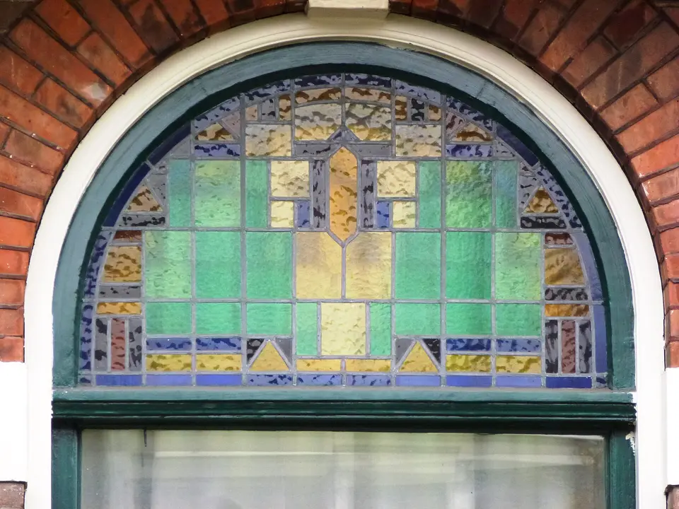 Een rond bovenlicht met glas-in-lood in groen, geel en blauw, in een groen geschilderd houten kozijn in een bakstenen gevel in Nijmegen